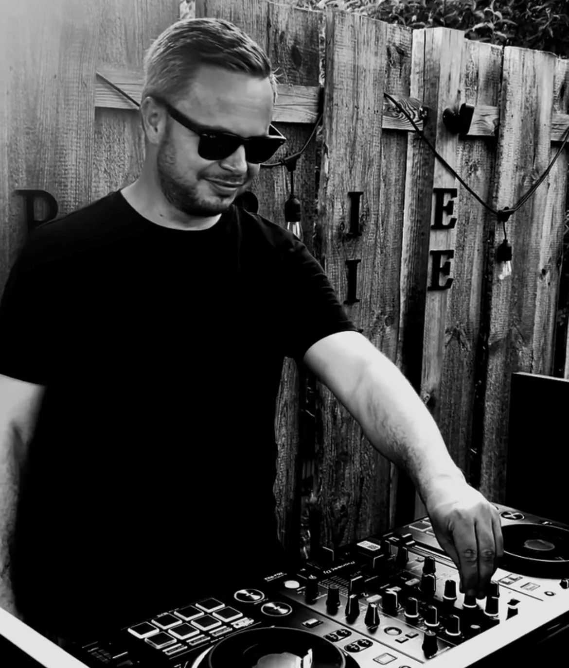 Me at my DJ controller in front of a wooden fence with string lights, smiling behind black sunglasses